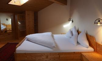 A cozy bedroom with a wooden bed and large pillows. The walls are light, and there is warm light that creates an inviting atmosphere.