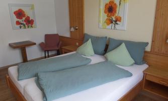 A cozy bedroom with a double bed and pillows in soft colors. On the wall, there are two pictures of flowers, and a small table and a chair are present in the room.