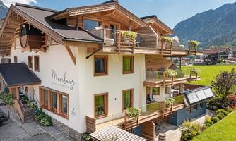 A modern building in alpine style with large windows and wood paneling. The surroundings are green and the mountains are visible in the background.