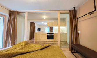 A modernly furnished bedroom with a gold-colored bed and a partition to the kitchen. The bright kitchen is well equipped and has large windows.