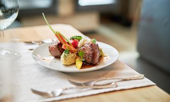 An elegant dish with juicy meat, colorful vegetables, and a side served on a white plate. The table is beautifully set with cutlery and a glass.