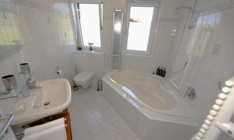A modern bathroom with white tiles and plenty of natural light. It features a bathtub, a shower, and a sink.