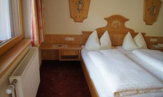 A cozy bedroom with two single beds and white cushions. The walls are in warm wood colors and there is a small chest of drawers as well as a window with curtains.