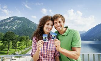 A couple is standing on a balcony holding tickets in front of the camera. In the background, green mountains and a clear lake can be seen.