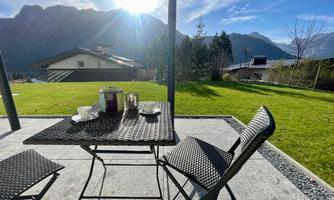 A cozy table with two chairs on a terrace. In the background, mountains and a clear sky can be seen.