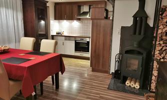 A cozy kitchen with a dining table and chairs. In the background, a wood stove and a cooking area can be seen.