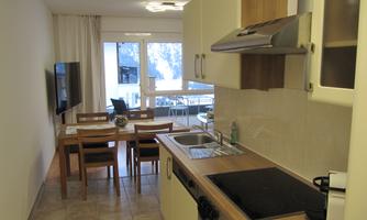 A modern kitchen with wooden cabinets and a dining table. Large windows provide plenty of light and a bright atmosphere.