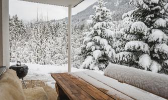 A cozy terrace with a wooden table, surrounded by snow-covered trees. In the background, mountains are visible in a winter landscape.