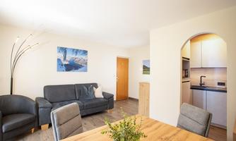 A bright living area with a cozy sofa and a dining table. The walls are decorated with modern pictures and the kitchen is visible in the background.