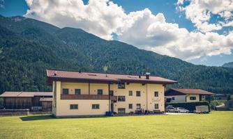 A large house in the mountains, surrounded by a green meadow. In the background, majestic mountains and a blue sky with white clouds can be seen.
