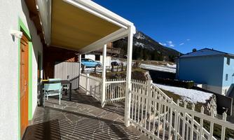 A cozy terrace with sun protection, chairs, and a table. In the background, snow-covered mountains and colorful houses can be seen.