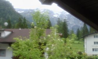 A view from a window onto a green landscape with trees and mountains in the background. The sky is cloudy, and there are some snow-capped peaks.
