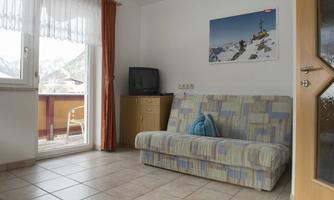 A simple living room with a sofa bed and a television. Bright light streams through the window and reveals a balcony.
