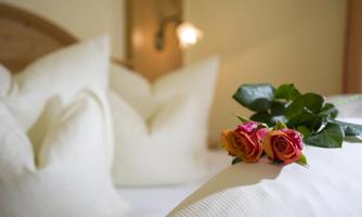 A cozy bedroom with white pillows and a wooden slice. Two roses with green leaves lie on the bed.