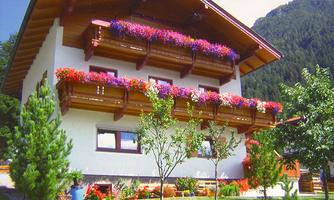 A beautiful house with balcony flowers and a green garden. In the background, mountains can be seen.