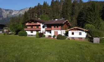 A charming wooden house surrounded by green meadows and trees. Majestic mountains are visible in the background.