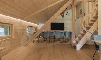 A modern, bright wooden room with a staircase and a long table. A television is mounted on the opposite wall, and large windows provide plenty of daylight.