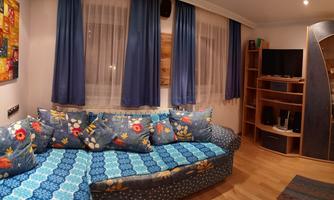 A cozy living room with a colorful couch and large windows. A vibrant painting hangs on the wall, and there is a bookshelf as well as a television.