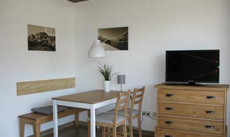 A bright room with a dining table, chairs, and a wooden dresser. Pictures hang on the wall, and there is a modern lamp as well as a television.