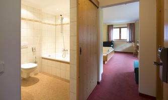 A modern bathroom with a bathtub and shower is visible. In the background, a cozy room with a window and television can be seen.
