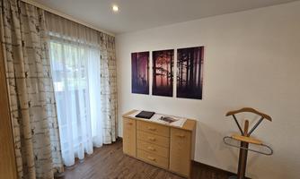 A cozy room with wooden flooring and large windows.  
On the wall hang pictures of a forest at sunset, and a wooden table stands in the middle.