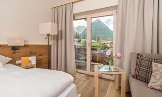 A cozy hotel room with a bright bed and a comfortable couch. Through the windows, you can see mountains and a lot of greenery.