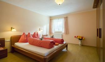 A bright bedroom with a double bed and red pillows. By the wall, there is a dresser and a vase with flowers.