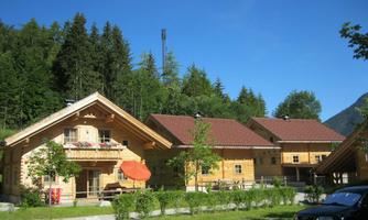 Two cozy wooden houses in an idyllic, green surroundings. In the background, there are tall trees and a clear blue sky.