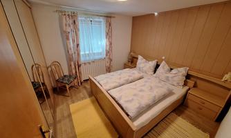 A cozy bedroom with a double bed and wooden paneling. There is a chair and a rug, with plenty of natural light from a window.