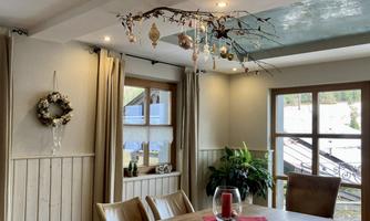 A cozy dining area with a wooden table and chairs. Festive decorations hang from the ceiling and a window offers a view outside.