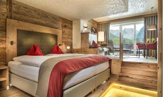 A modern hotel room with wooden panels and a large double bed. The room provides access to a balcony with a view of the mountains.