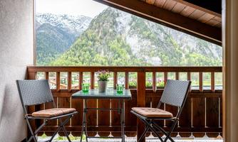 A beautiful balcony with two chairs and a small table. In the background, mountains with green forests can be seen.