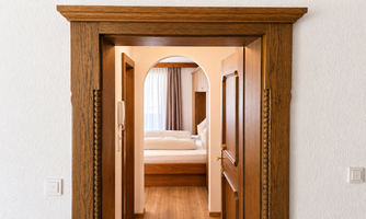 An entrance with a wooden door frame, leading to a bright bedroom. In the background, one can see a bed and windows with curtains.
