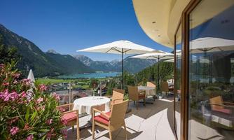 A quiet terrace overlooking a clear lake and the surrounding mountains.  
Umbrellas and cozy seating invite relaxation.