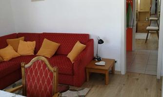 A cozy living room with a red sofa and yellow cushions. In the background, a table and access to another room can be seen.