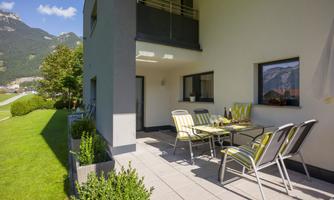 A modern terrace with a dining table and chairs. In the background, you can see a green meadow and mountains.