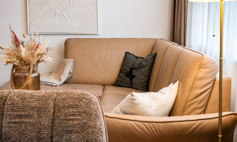 A cozy living room with a beige leather sofa and a decorative pillow. Next to the sofa stands a lamp and a table with a vase full of dried flowers.