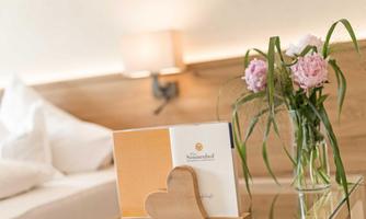 A cozy bedroom with a wooden heart and a vase of pink flowers. On the nightstand lies a book with a logo.