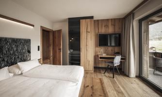A modern room with wooden walls and a double bed. It features a desk, a television, and large windows overlooking nature.