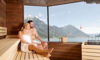 A couple relaxes in a sauna with a view of the mountains and a lake. The wooden paneling and the large window create a cozy atmosphere.