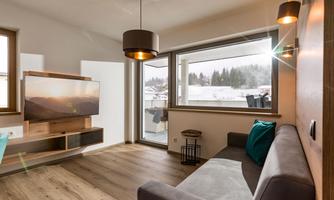 A modern living room with a gray sofa and a television on the wall. Large windows let in plenty of daylight and offer a view of the surrounding nature.