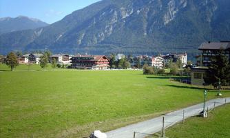 A tranquil landscape with a vast green field and mountains in the background. In the foreground, some buildings and a path are visible.