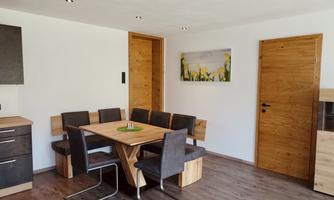 A modern dining area with a large wooden table and comfortable chairs. The walls are painted light, and there is a door as well as a window for plenty of daylight.