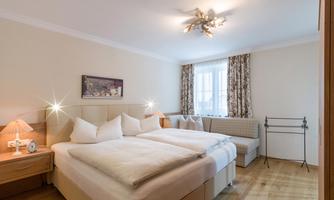 A bright bedroom with two single beds and wooden flooring. The walls are painted in soft colors, and there are windows with curtains as well as a cozy seating area.