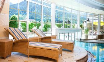 An elegant interior with two loungers next to a pool. Large windows provide a view of the mountains and plenty of daylight.