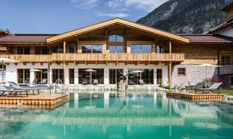 A modern building with wooden cladding and large windows. The pool in the foreground reflects the surrounding mountains.