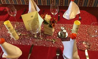 An elegantly set table with red tablecloths, glasses, and decorated napkins. In the center, there is a flower arrangement and a card for a special occasion.