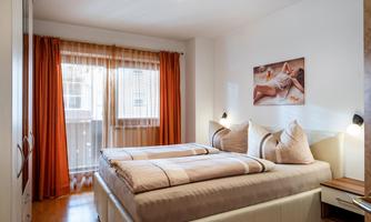 A modern bedroom with a double bed and soft textiles. Large windows with orange curtains create a bright and inviting atmosphere.