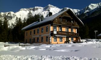 A cozy house in the snow with an impressive mountain landscape in the background. The sky is clear and blue.
