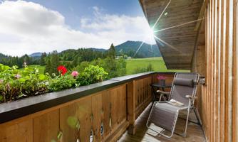 A cozy balcony with a relaxing lounger and blooming plants. In the background, a green landscape stretches under a clear sky.